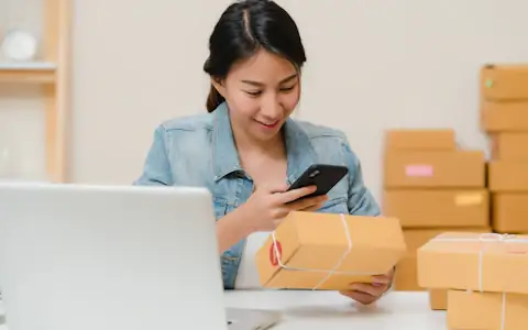 Online seller checking a parcel with her phone beside a laptop and boxes, cross-border e-commerce