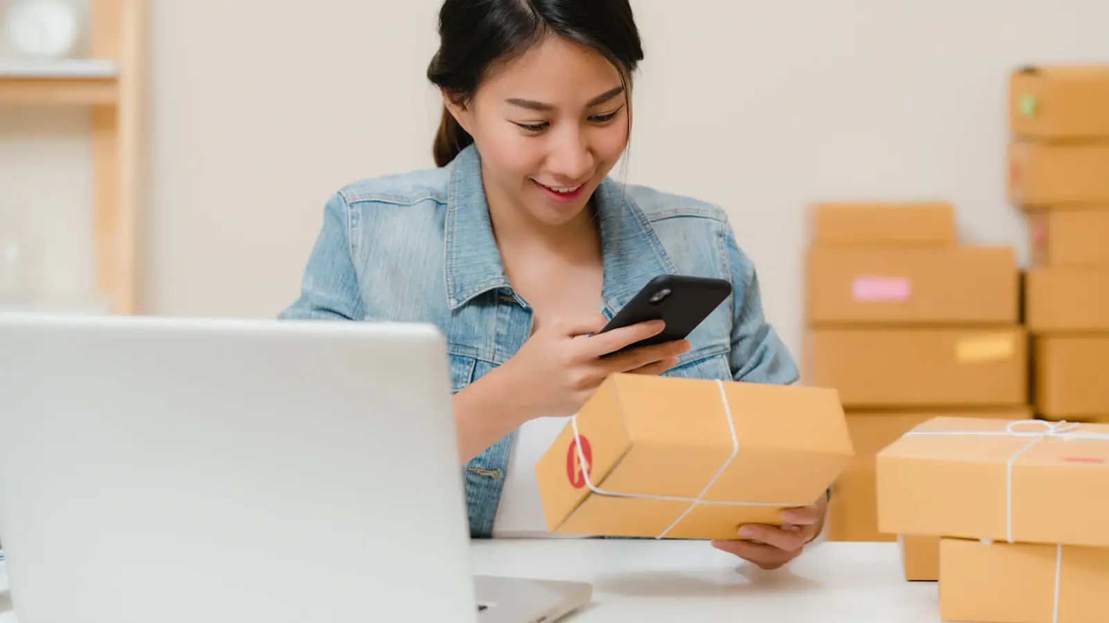 Online seller checking a parcel with her phone beside a laptop and boxes, cross-border e-commerce
