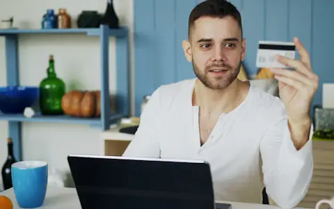 Man holding a payment card at his laptop in a bright kitchen, paying online