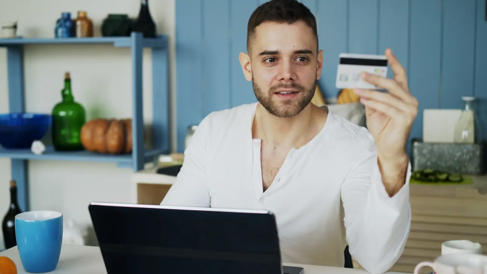 Man holding a payment card at his laptop in a bright kitchen, paying online