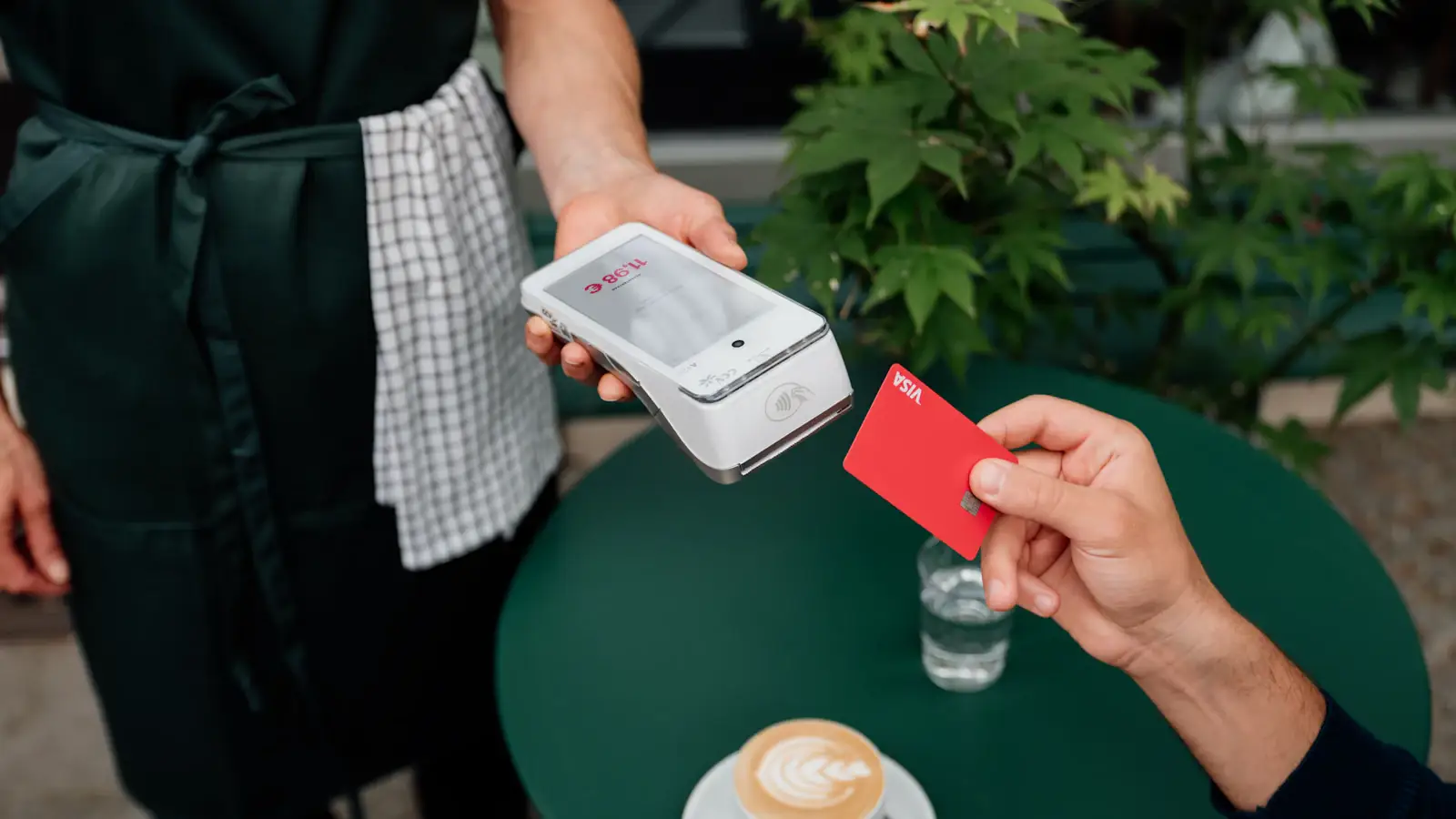 Customer tapping a Visa card on a payment terminal at a cafe table, a card network settlement in action